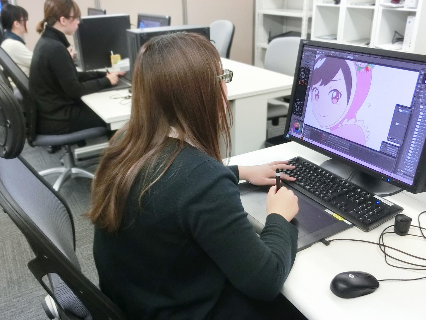 A female animator in the foreground sits facing away from the camera at a computer. She is drawing on a digital pad with a stylus in her right hand, and a color drawing is displayed on the monitor screen. Her left hand is hovering on a keyboard. A female animator in the foreground sits facing away from the camera at a computer. She is drawing on a digital pad with a stylus in her right hand, and a color drawing is displayed on the monitor screen. Her left hand is hovering on a keyboard.