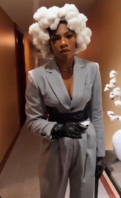 Serena Williams wearing a gray suit with a deep V-neck jacket, black belt, and black gloves. She has a voluminous white cloud-like hairstyle. The background shows a hallway with beige walls, a carpeted floor, and decorative white objects. Serena Williams wearing a gray suit with a deep V-neck jacket, black belt, and black gloves. She has a voluminous white cloud-like hairstyle. The background shows a hallway with beige walls, a carpeted floor, and decorative white objects.