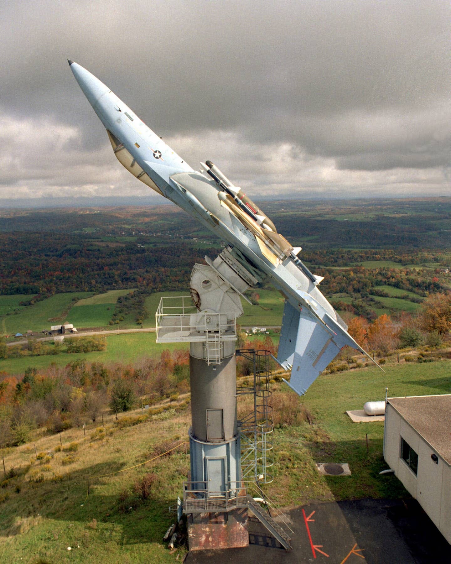 A right side view of an F-15C Eagle aircraft mounted upside-down on a pedestal at the Rome Air Development Center's Newport test site, October 1988. A radar warning system pod mounted on the fuselage is being evaluated in comparison with the aircraft's onboard radar warning system. <em>National Archives</em>
