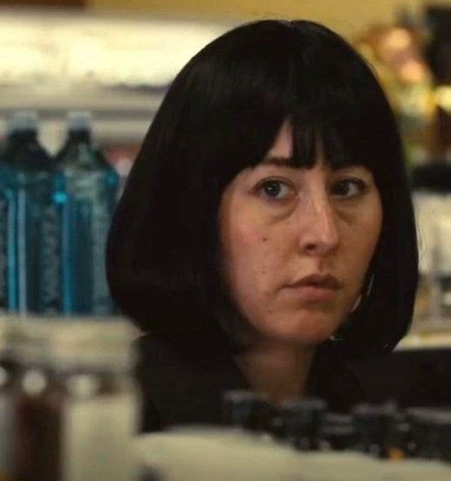 A woman with short, dark bobbed hair, wearing a dark jacket, standing in a store aisle. Bottles of water and other items are visible on shelves behind her.