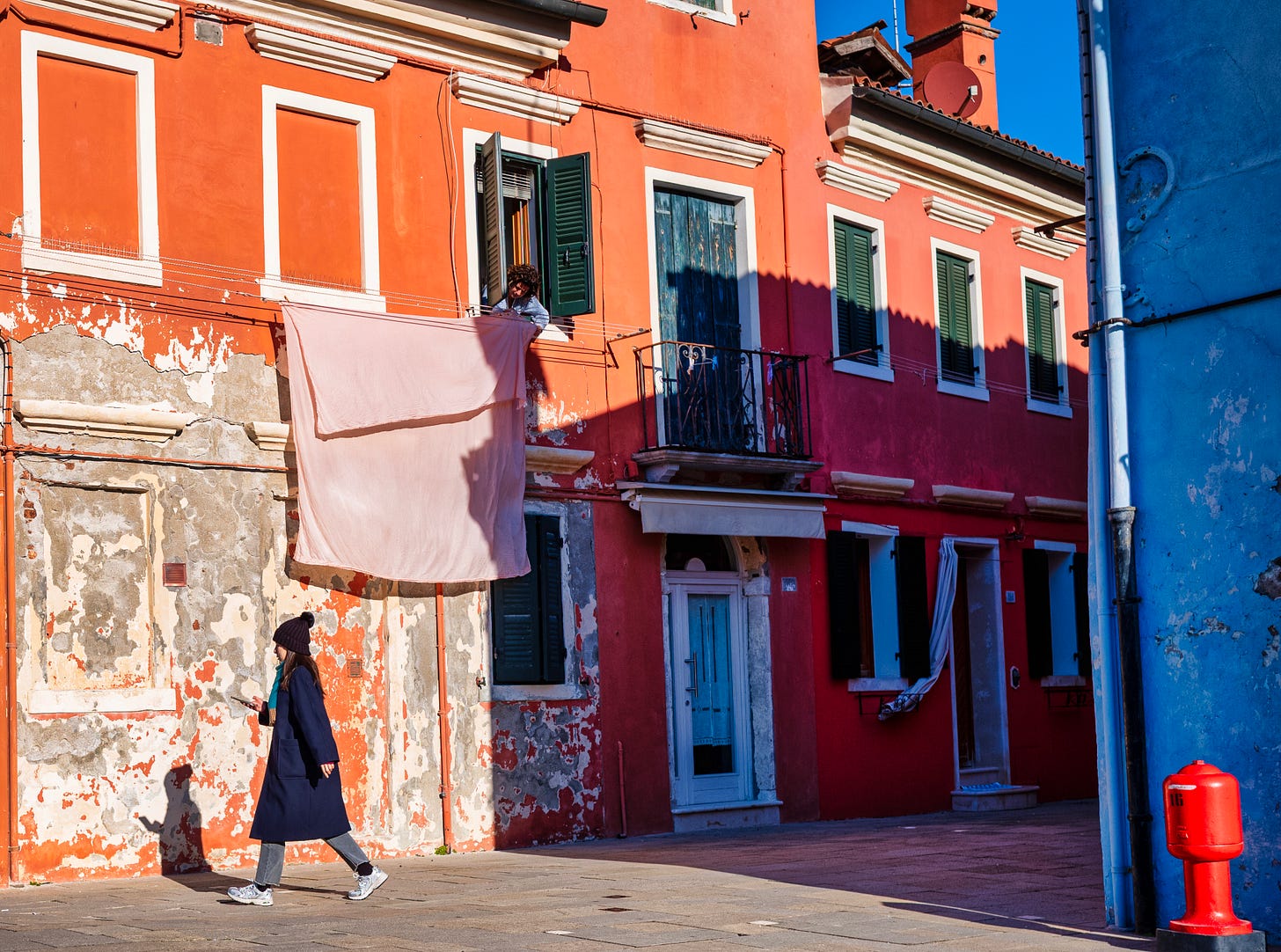 A woman walks past a weathered red house in Burano while another hangs laundry from an upper window, both lit by crisp winter light.