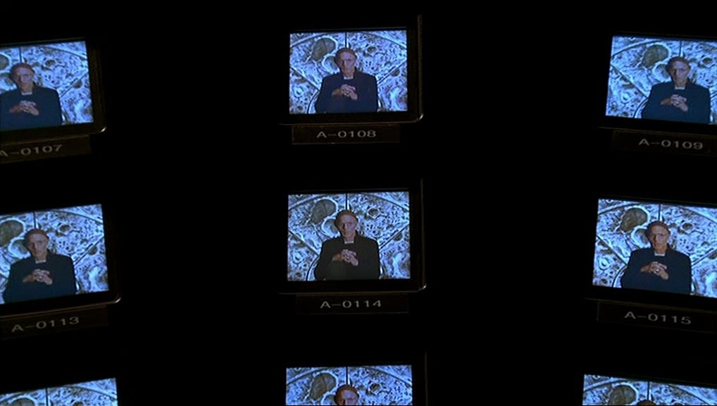 Several small surveillance screens display the same man speaking, arranged in a dark grid that emphasizes constant monitoring.