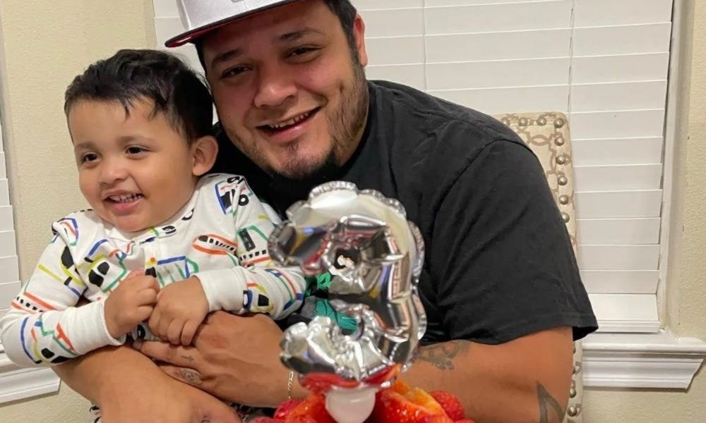 color photo of Kilmar Abrego Garcia, wearing a black T-shirt and baseball cap, holding his giggling son in his arms. The boy is wearing pajamas. An inflatable mylar table decoration in the foreground with the numeral '3' makes clear it's the boy's birthday color photo of Kilmar Abrego Garcia, wearing a black T-shirt and baseball cap, holding his giggling son in his arms. The boy is wearing pajamas. An inflatable mylar table decoration in the foreground with the numeral '3' makes clear it's the boy's birthday