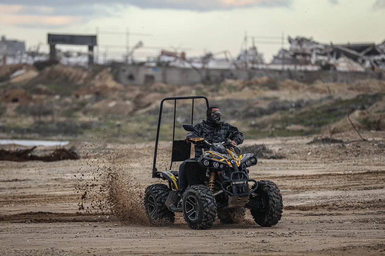 오프로드용 사륜차량이 모래밭을 달리고 있다. 뒤로는 폐허인 듯한 마을이 흐릿하게 보인다.