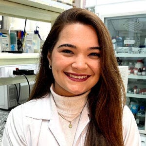 Mulher branca, cabelos longos pretos. Ela usa um jaleco branco por cima de uma blusa de gola branca e está sentada dentro de um laboratório. Mulher branca, cabelos longos pretos. Ela usa um jaleco branco por cima de uma blusa de gola branca e está sentada dentro de um laboratório.