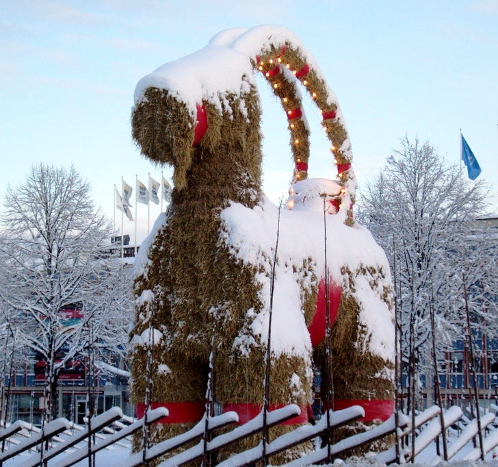 a large snow-covered straw goat stands in the foreground. The goat is decorated with red ribbon and white lights. There is a wooden lattice fence around the goat. The background is snow-covered trees and some local flags.