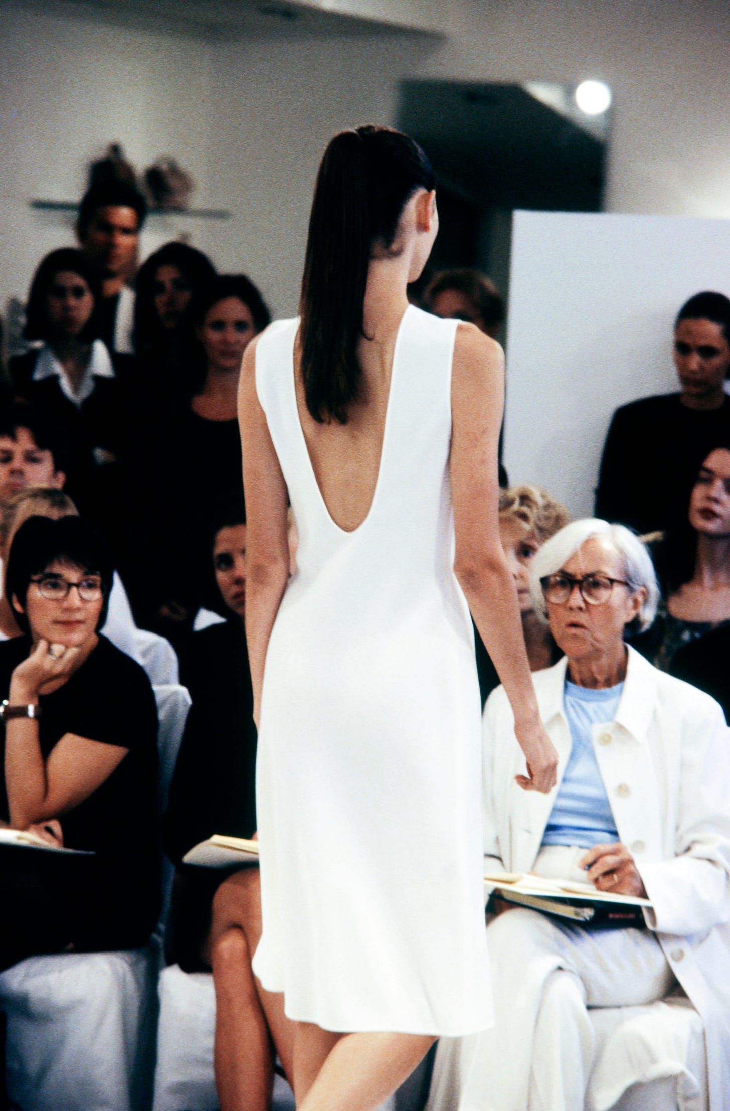 A seated woman with cropped silver hair and wearing a white ensemble looks on as a model clad in a minimal white dress walks down the runway.