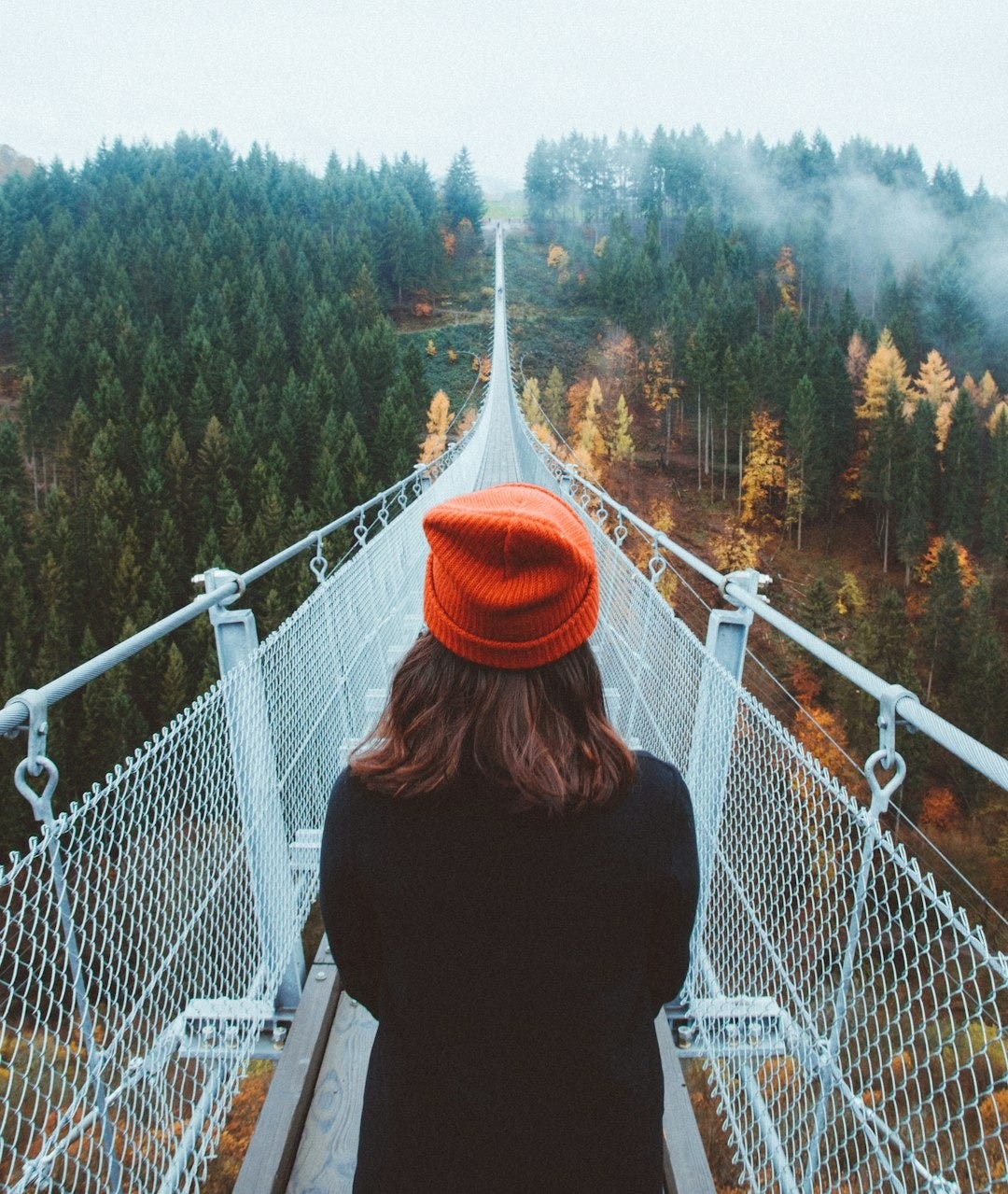woman wearing knit cap walking on white bridge between trees during daytime