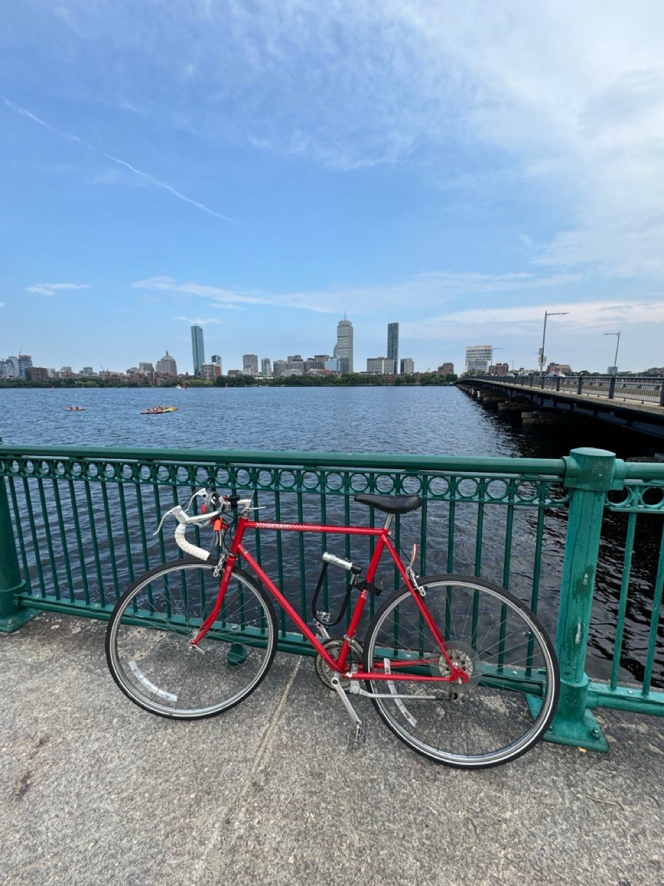A red Peugeot road bike leaning against a green railing beside the Charles River, with the Boston skyline and Harvard Bridge behind it A red Peugeot road bike leaning against a green railing beside the Charles River, with the Boston skyline and Harvard Bridge behind it