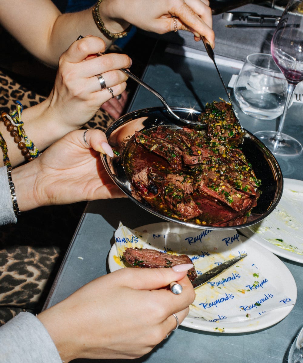 A group of hands diving into a plate of steak with gremolata on top, with a gray table with wine glasses next to it