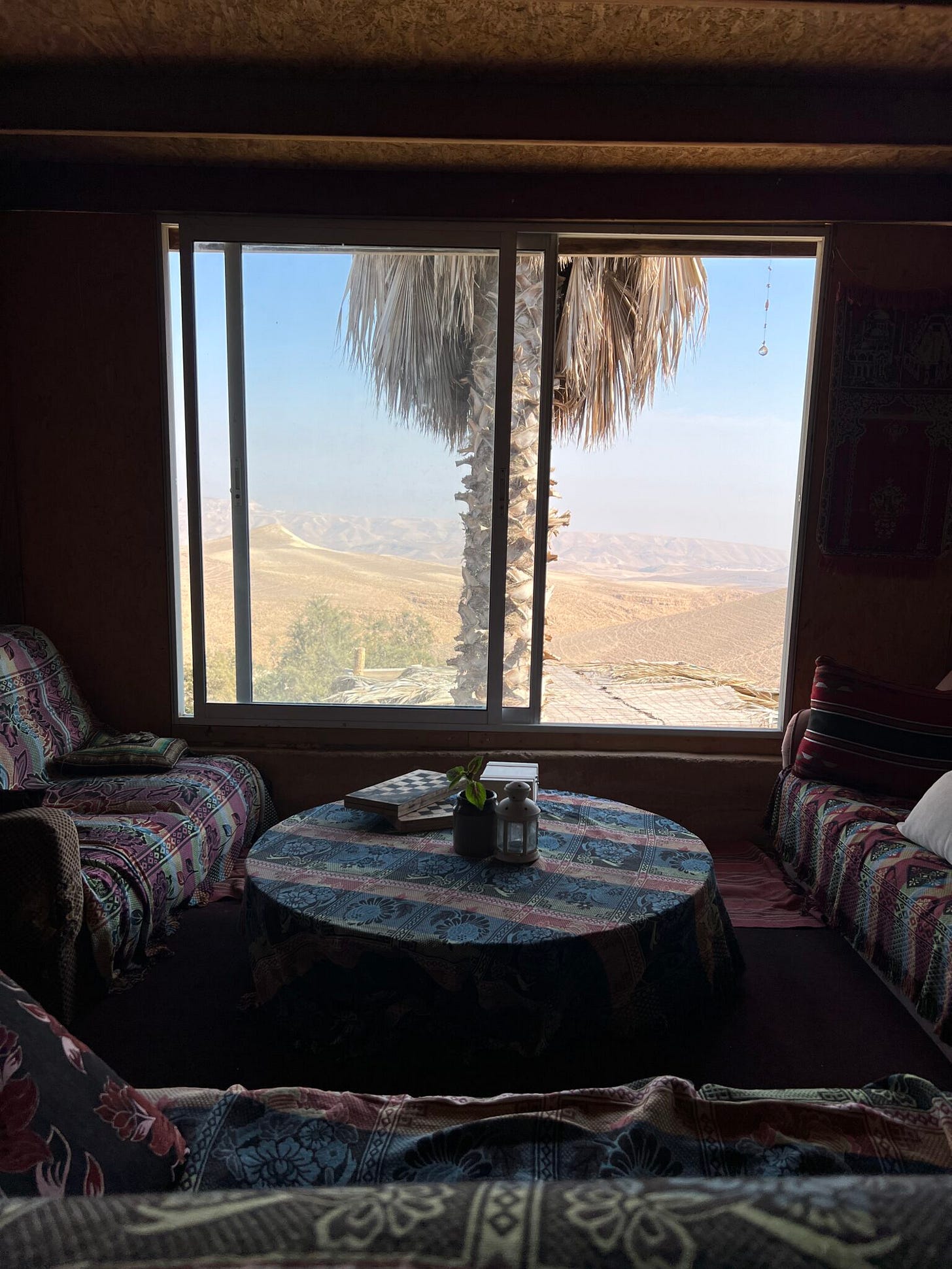 Die Bleibe eines Aussteigers, der in Arad Retreats anbietet. Ein gemütlicher Raum mit sofas und rundem Tisch in der Mitte, ein großes Fenster mit Palme davor und Blick auf die Wüste Die Bleibe eines Aussteigers, der in Arad Retreats anbietet. Ein gemütlicher Raum mit sofas und rundem Tisch in der Mitte, ein großes Fenster mit Palme davor und Blick auf die Wüste