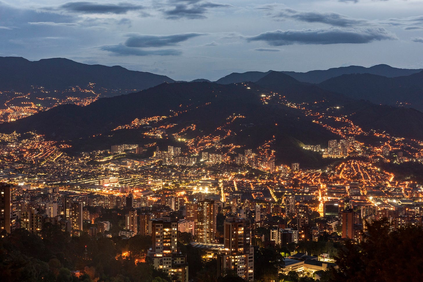 Medellín, Colombia at dusk — city lights filling the valley floor and climbing the surrounding Andean hillsides, with lit apartment towers in the foreground.