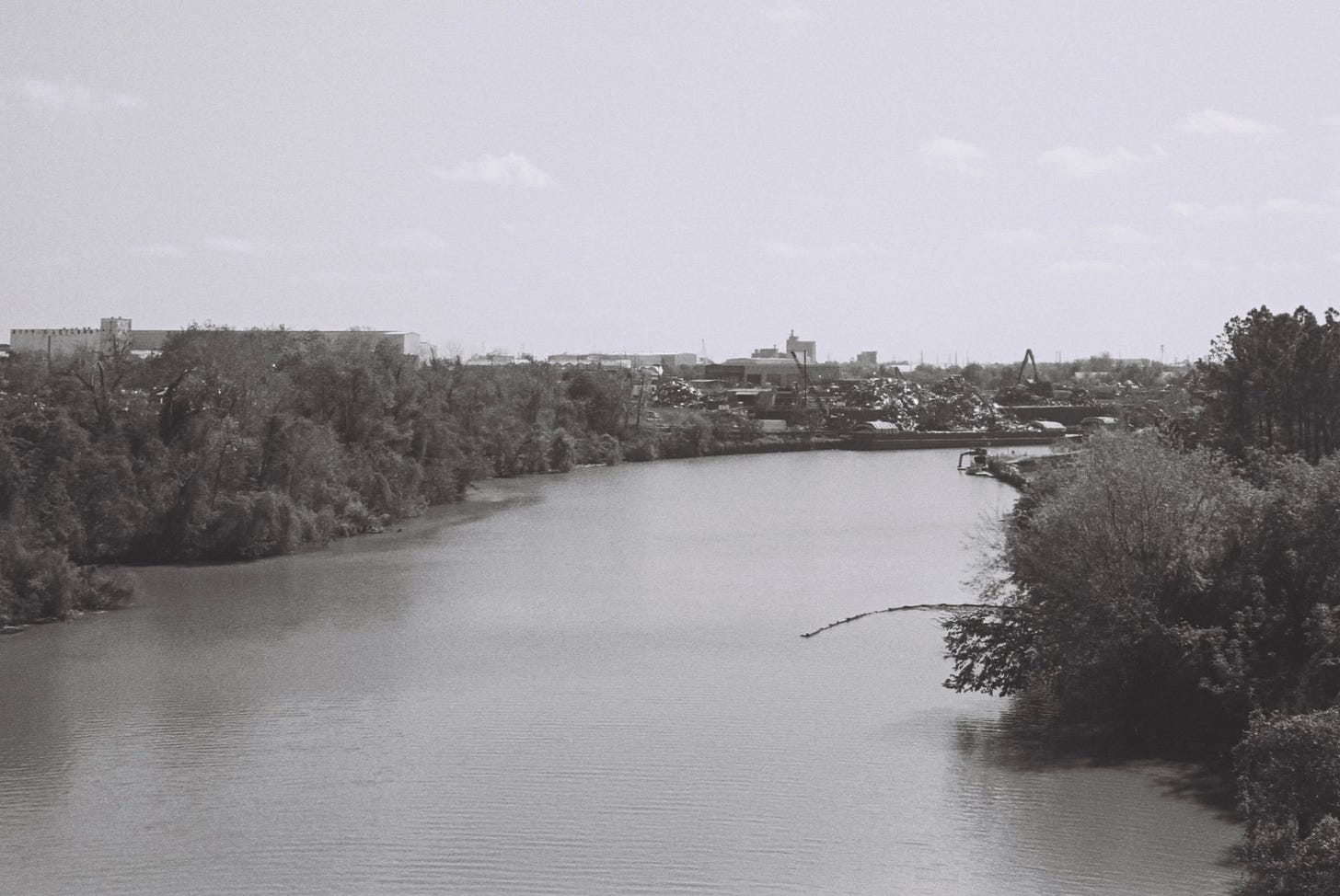 Black and white photo of river with tree-lined banks and scrap metal yard on one section Black and white photo of river with tree-lined banks and scrap metal yard on one section