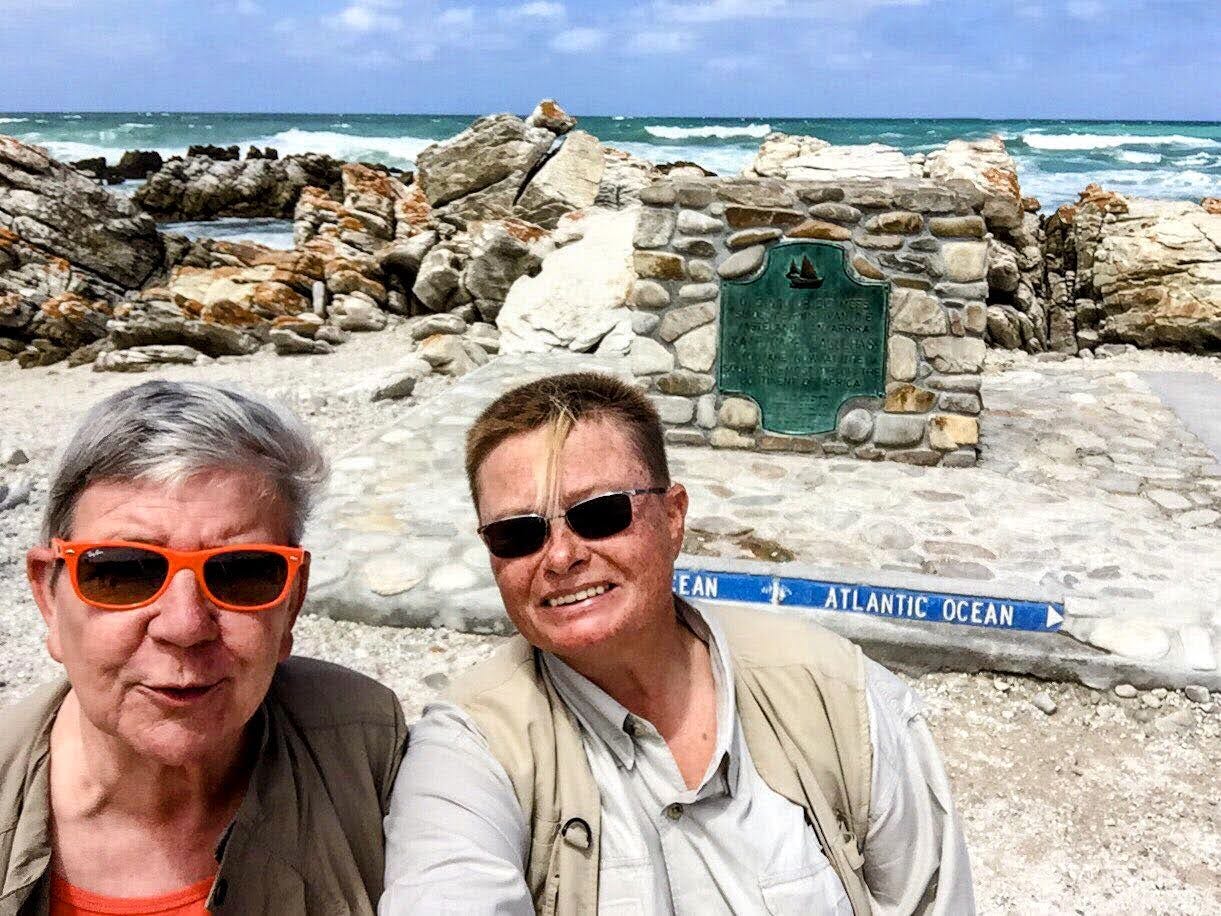 Two people wearing sunglasses and beige outdoor vests smile for a selfie at Cape Agulhas, South Africa. Behind them, rocky shoreline and a green plaque mark the point where the Indian and Atlantic Oceans meet. Waves crash in the background under a bright blue sky.