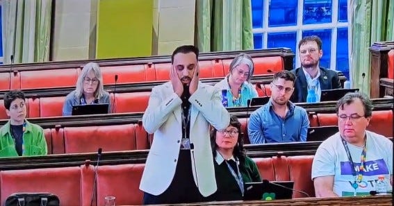 Councillor Abdul Malik standing in Bristol City Council's Full Council chamber, hands pressed against his ears, eyes closed, performing the Adhan — the Islamic Call to Prayer — during the public forum on 10th March 2026. Councillors and members of the public are visible seated around him in the red-seated chamber -https://bit.ly/4upw6aP