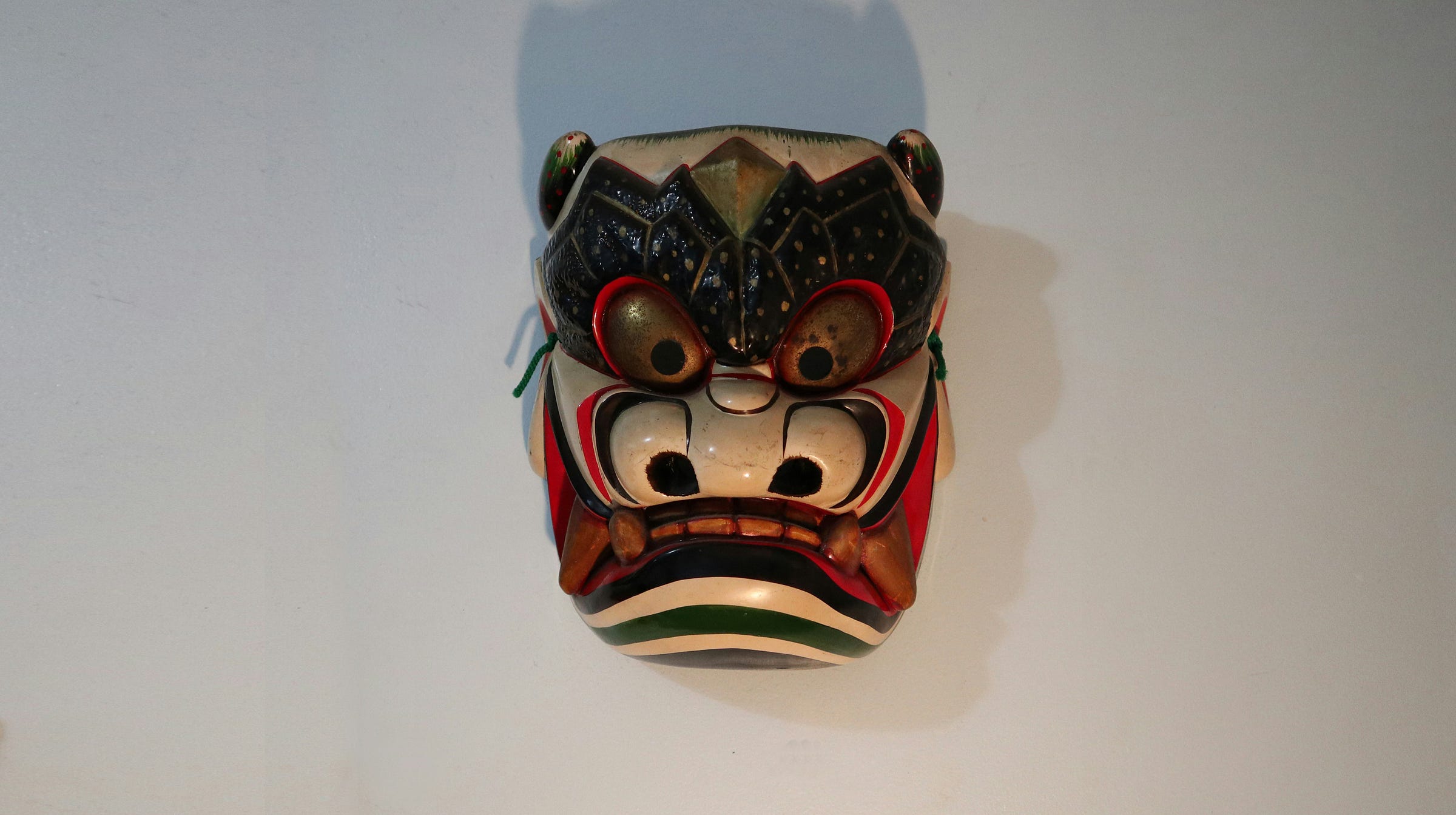 A traditional, hand-painted mask with exaggerated features, including large, bulging eyes, sharp teeth, and intricate patterns, mounted on a plain wall. A traditional, hand-painted mask with exaggerated features, including large, bulging eyes, sharp teeth, and intricate patterns, mounted on a plain wall.