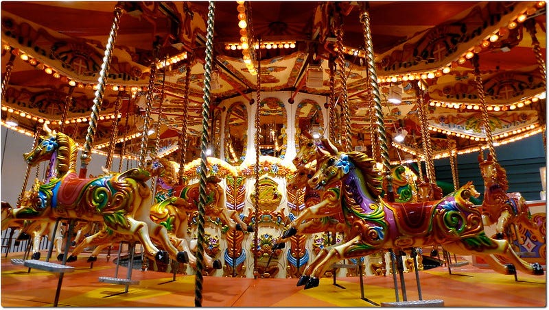 A carousel stands, brightly light, the poles and horses painted in gold, green, blue, pink, and red A carousel stands, brightly light, the poles and horses painted in gold, green, blue, pink, and red