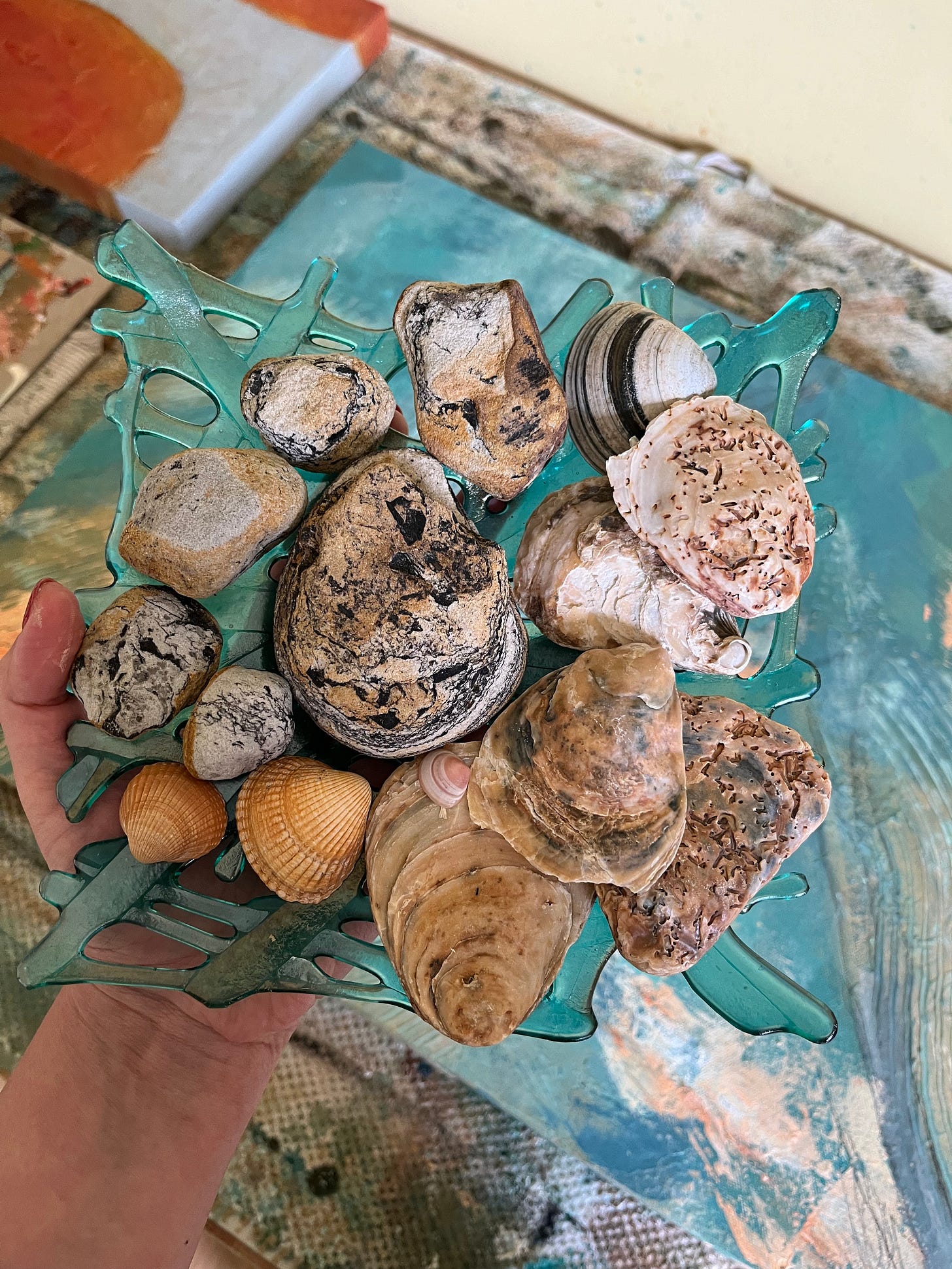 A hand holding a glass dish filled with collected seashells and stones, each with unique textures and colors. The dish has an organic, seaweed-like design in translucent teal. In the background, abstract paintings in oceanic hues of blue and earthy tones rest on a paint-stained surface, blending art and nature in a quiet, reflective moment