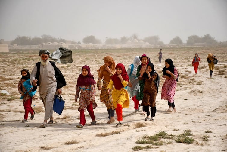 An older man in a turban leads a group of young girls in colorful dresses and headscarves along a dusty terrain.