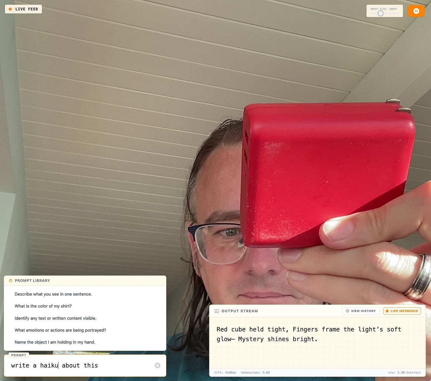 Screenshot of a man with glasses holding a red cube-shaped object up to the camera in a live computer vision interface; top left label reads “LIVE FEED”; top right slider label reads “INPUT SIZE: 480PX”; lower left panel titled “PROMPT LIBRARY” with prompts “Describe what you see in one sentence.” “What is the color of my shirt?” “Identify any text or written content visible.” “What emotions or actions are being portrayed?” “Name the object I am holding in my hand.”; below that a field labeled “PROMPT” containing the text “write a haiku about this”; lower right panel titled “OUTPUT STREAM” with buttons “VIEW HISTORY” and “LIVE INFERENCE” and generated text “Red cube held tight, Fingers frame the light’s soft glow– Mystery shines bright.”; a small status bar at the bottom shows “ttft: 4188ms tokens/sec: 5.09” and “ctx: 3.3B-Instruct”. Screenshot of a man with glasses holding a red cube-shaped object up to the camera in a live computer vision interface; top left label reads “LIVE FEED”; top right slider label reads “INPUT SIZE: 480PX”; lower left panel titled “PROMPT LIBRARY” with prompts “Describe what you see in one sentence.” “What is the color of my shirt?” “Identify any text or written content visible.” “What emotions or actions are being portrayed?” “Name the object I am holding in my hand.”; below that a field labeled “PROMPT” containing the text “write a haiku about this”; lower right panel titled “OUTPUT STREAM” with buttons “VIEW HISTORY” and “LIVE INFERENCE” and generated text “Red cube held tight, Fingers frame the light’s soft glow– Mystery shines bright.”; a small status bar at the bottom shows “ttft: 4188ms tokens/sec: 5.09” and “ctx: 3.3B-Instruct”.