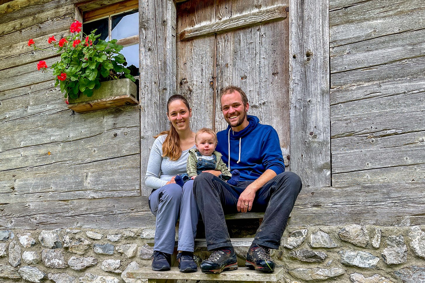 Eine junge Bauernfamilie sitzt während der Alpzeit auf den Steinstufen vor der wettergegerbten Holztür einer traditionellen Alphütte. Links blüht ein roter Geranienkasten unter einem kleinen Fenster. Die Eltern, beide lächelnd, tragen Outdoor-Kleidung, der Vater in dunkler Hose und blauem Kapuzenpulli, die Mutter in grauem Langarmshirt. Ihr Baby sitzt zwischen ihnen, warm eingepackt in kariertem Hemd und Latzhose. Die rustikale Holzhütte mit Natursteinfundament betont die alpine Umgebung. Eine junge Bauernfamilie sitzt während der Alpzeit auf den Steinstufen vor der wettergegerbten Holztür einer traditionellen Alphütte. Links blüht ein roter Geranienkasten unter einem kleinen Fenster. Die Eltern, beide lächelnd, tragen Outdoor-Kleidung, der Vater in dunkler Hose und blauem Kapuzenpulli, die Mutter in grauem Langarmshirt. Ihr Baby sitzt zwischen ihnen, warm eingepackt in kariertem Hemd und Latzhose. Die rustikale Holzhütte mit Natursteinfundament betont die alpine Umgebung.