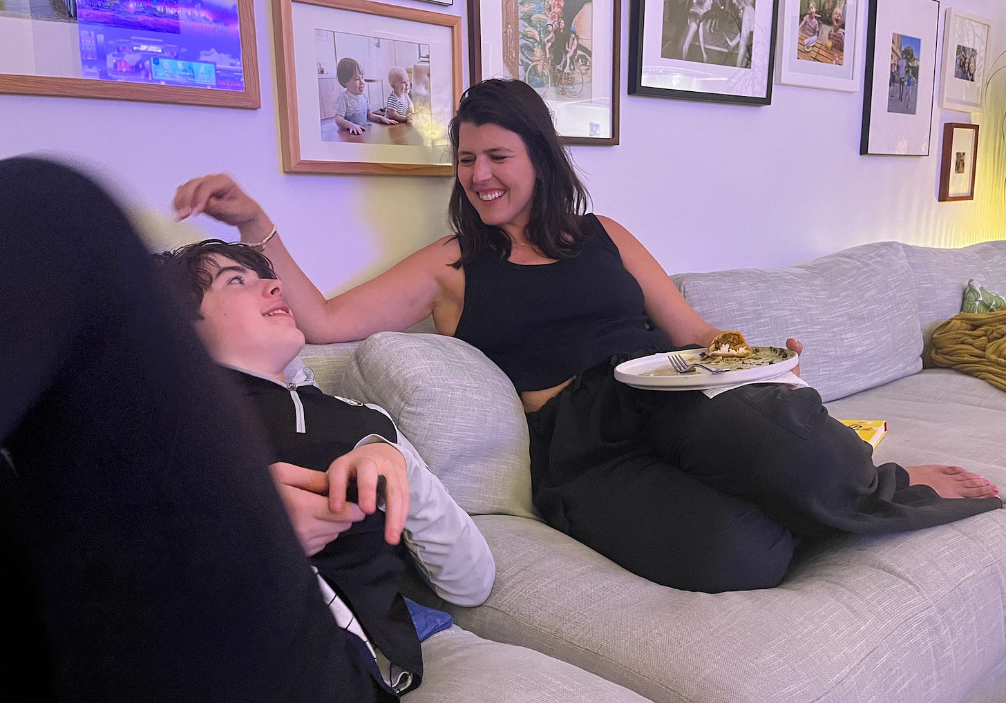 A smiling woman in a black sleeveless top sits on a gray couch with a plate of food, playfully touching the hair of a teenager who is reclining next to her. The teen, wearing a black hoodie, looks up at her with a relaxed expression. Framed photos decorate the walls behind them.