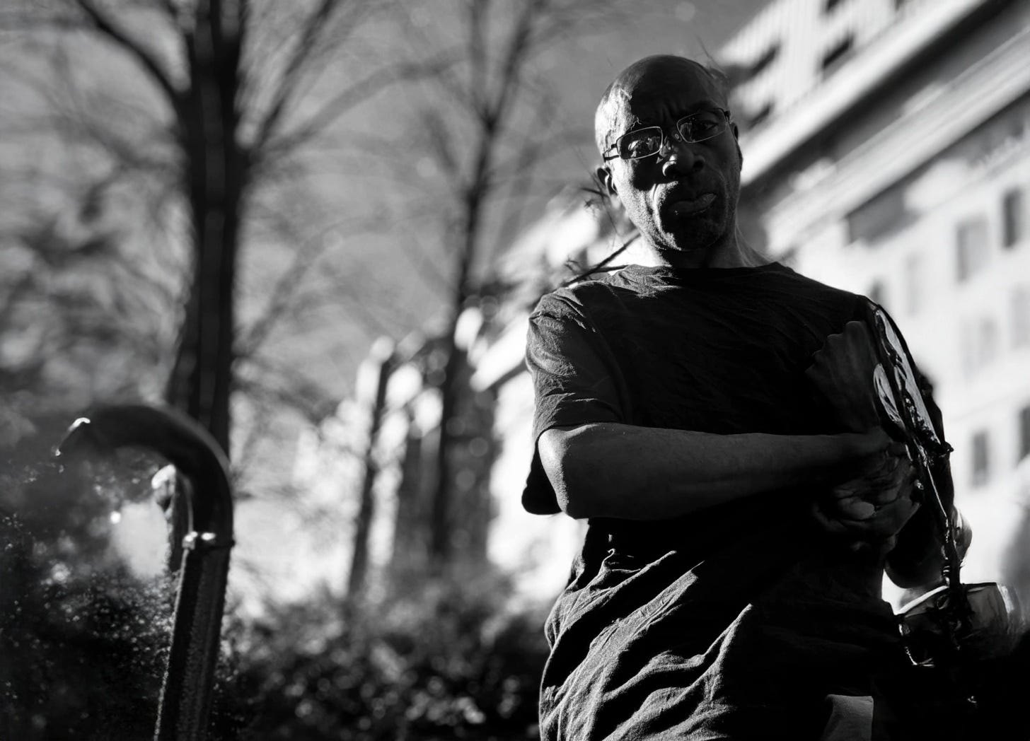 A black and white photograph of a Black man seated in a t-shirt, arms folded, with a tree and building in the background blurred.