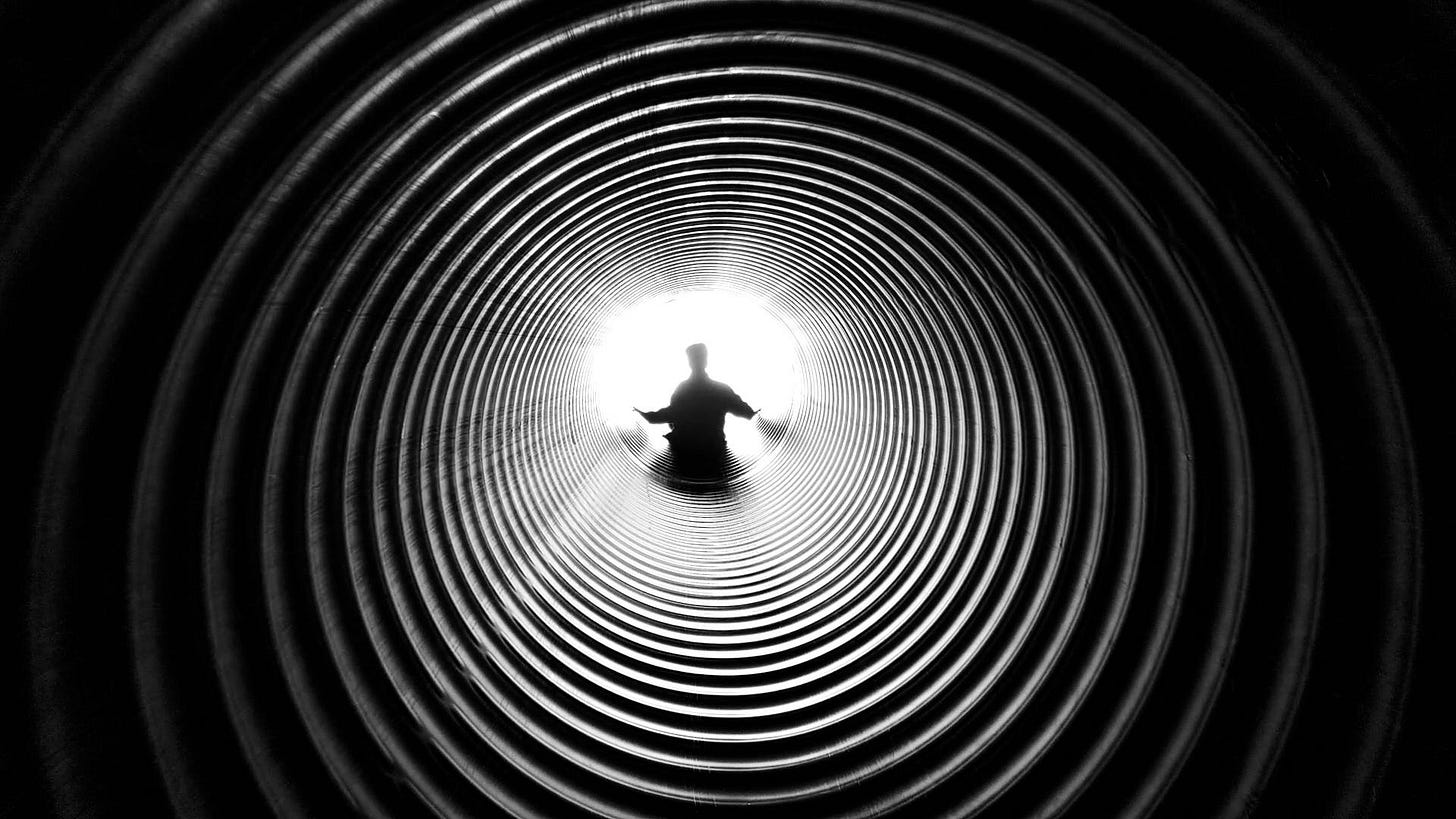 Black and white photo of a silhouetted figure sitting at the end of a tunnel lined with concentric rings, light pouring in behind them.