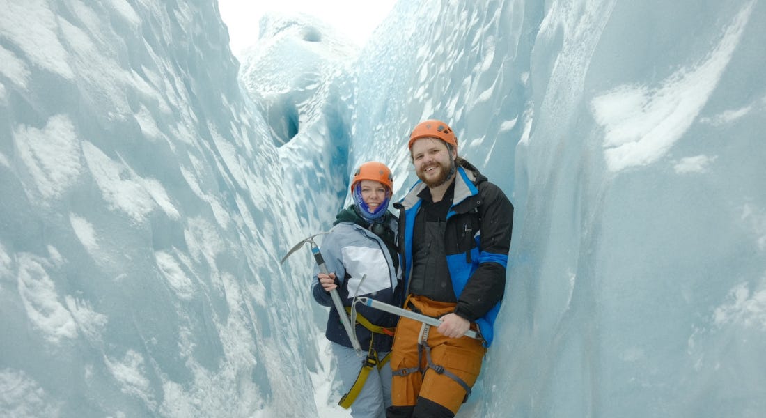 Two people wearing orange helmets and snow gear standing in the middle of three snowy peaks