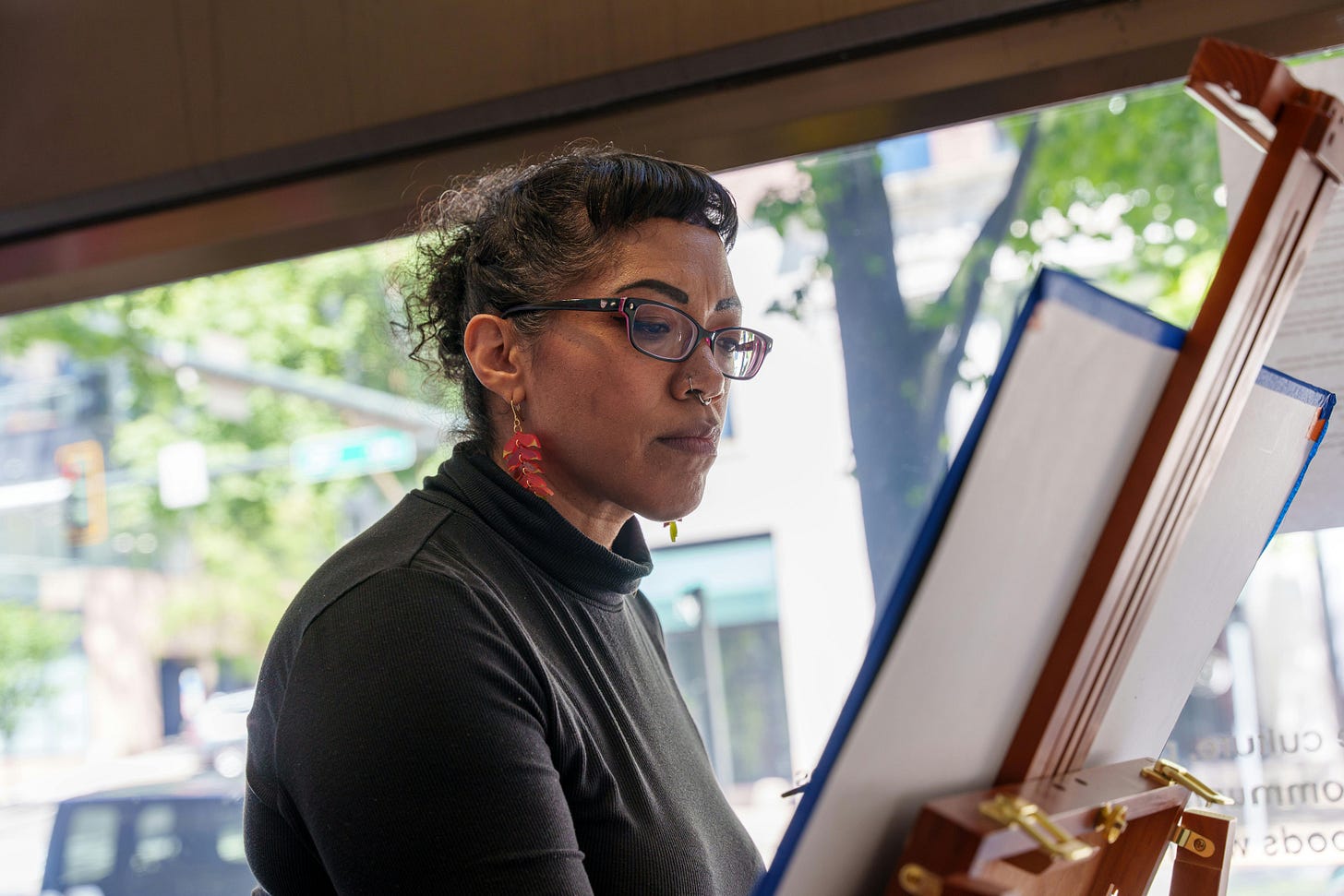 A middle-aged Black woman with her hair pinned up wears a black turtleneck, red drop earrings and black glasses as she paints a canvas on an easel beside a large window. A middle-aged Black woman with her hair pinned up wears a black turtleneck, red drop earrings and black glasses as she paints a canvas on an easel beside a large window.