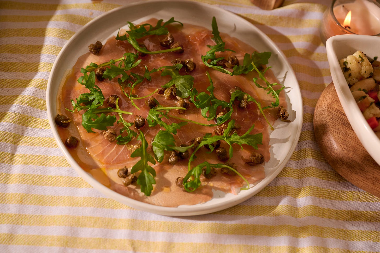 Plate of tuna carpaccio topped with fried capers and fresh arugula, drizzled with olive oil and lemon, served on a striped yellow and white tablecloth.