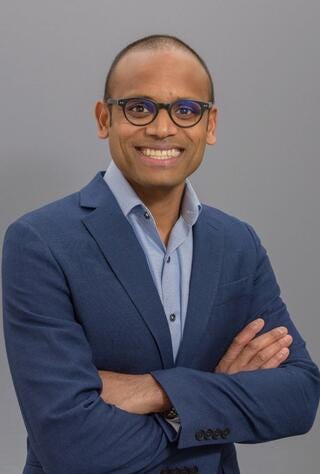 A Black man with dark eyes and black rimmed glasses smiles at the camera. He has little hair on his head and is clean shaven. He is wearing a dark blue blazer and a light blue shirt. 