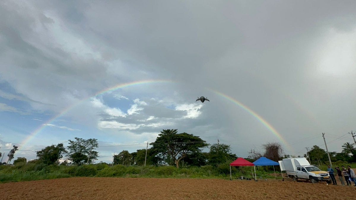 A rainbow arches across a cloudy sky with patches of blue. A bird is flying near the rainbow\'s center. Below, a field of plowed earth stretches toward trees and vegetation. Tents, a white van, and people are visible on the right side.