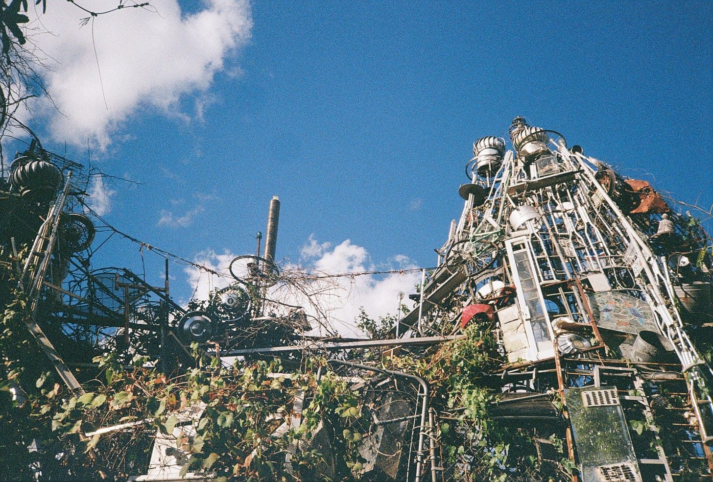 A photo shot from the ground and toward the bright blue sky featuring one side of the Cathedral of Junk. The tower reaches about two stories, leaves and vines winding through it.
