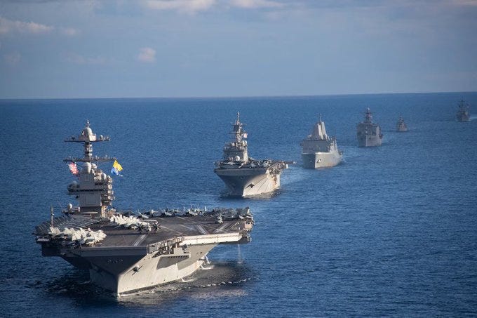 Aerial view over calm blue ocean showing multiple gray U.S. Navy warships in formation including aircraft carrier with angled flight deck and multiple destroyers with radar masts and missile launchers sailing in line under partly cloudy sky.
