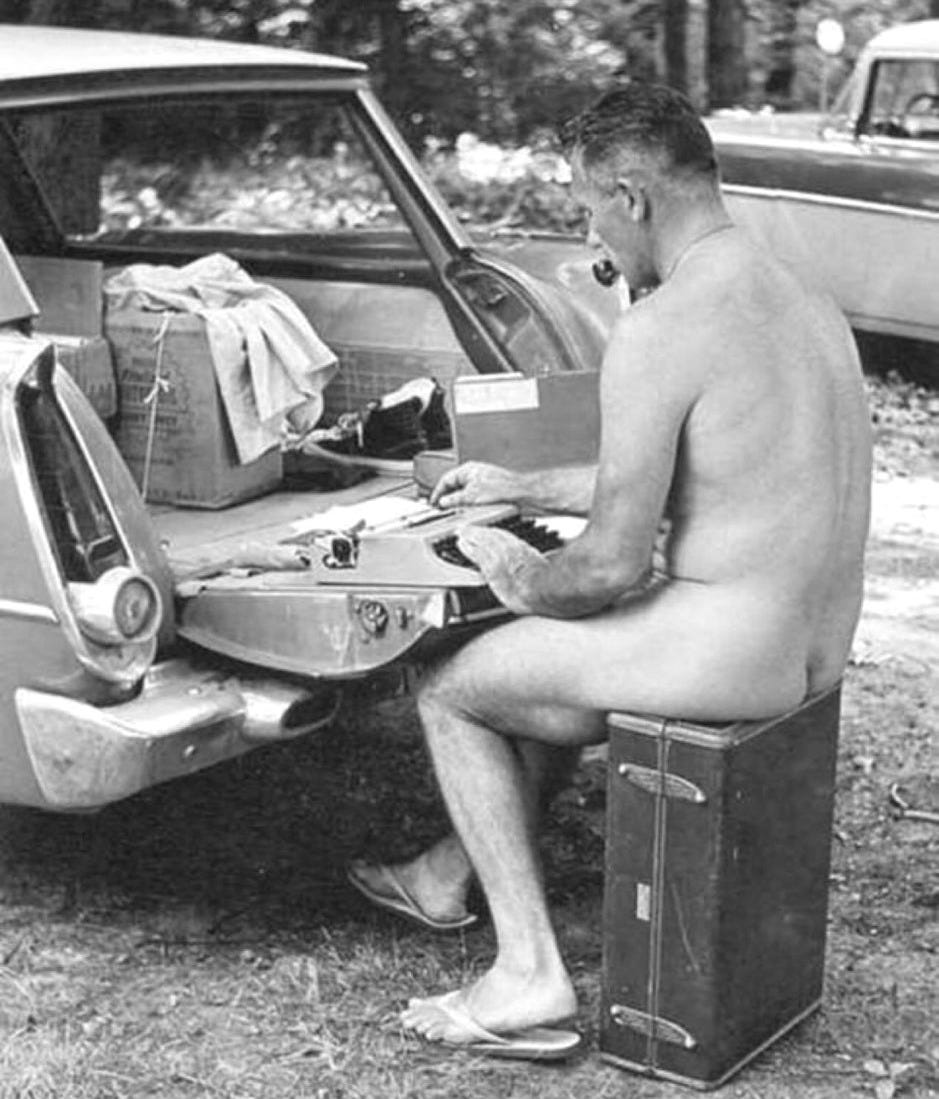 A black-and-white photograph of a nude man seated on a suitcase behind an open car trunk, writing at a portable typewriter outdoors. Papers, boxes, and clothing are visible in the trunk. The scene appears informal and workmanlike, with parked cars and trees in the background.