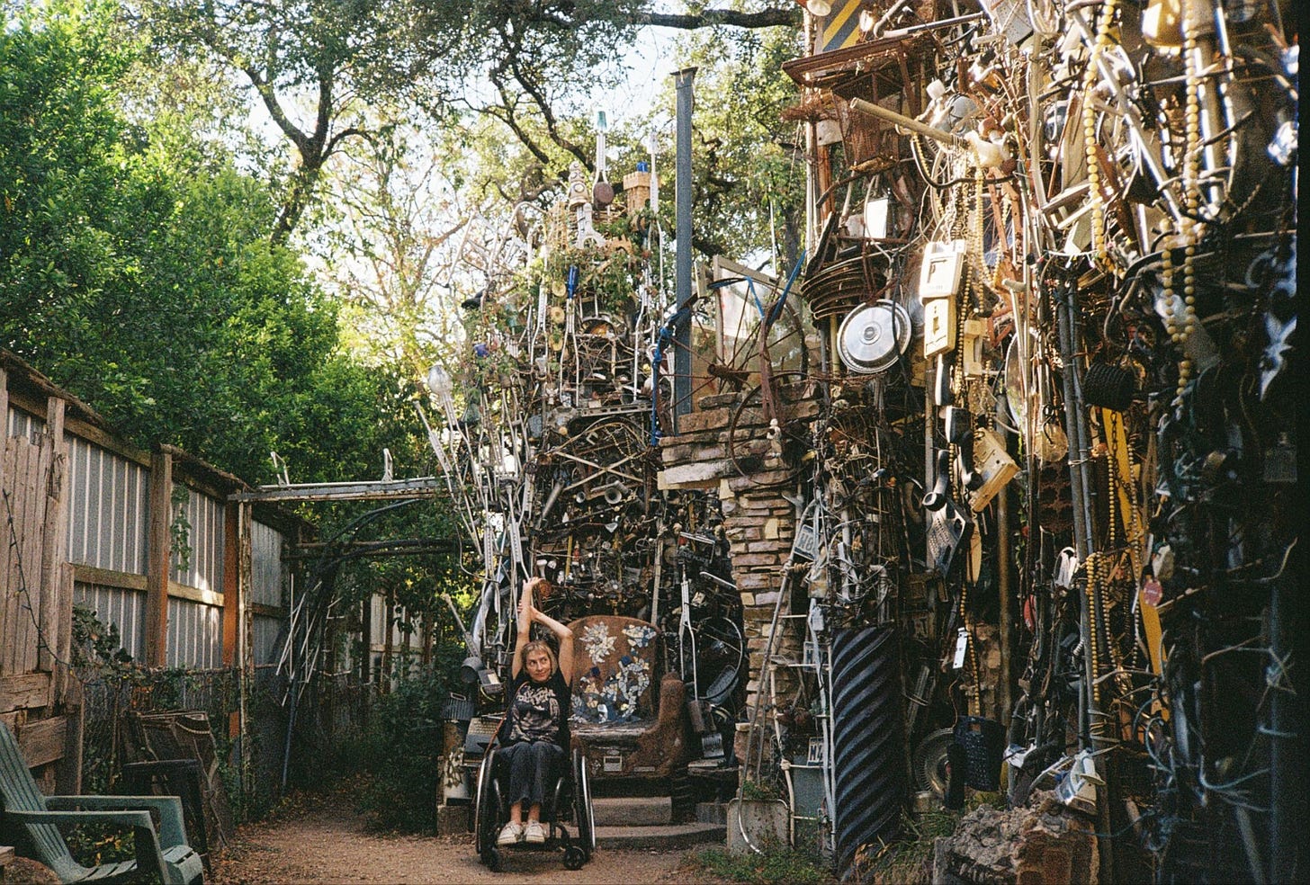 A photo of Rebekah sitting in front of the mosaic throne, a fence and trees to her right, the Cathedral of Junk towering to her left. Her arms are reaching toward the sky as she eyes the tangled, glimmering tower.