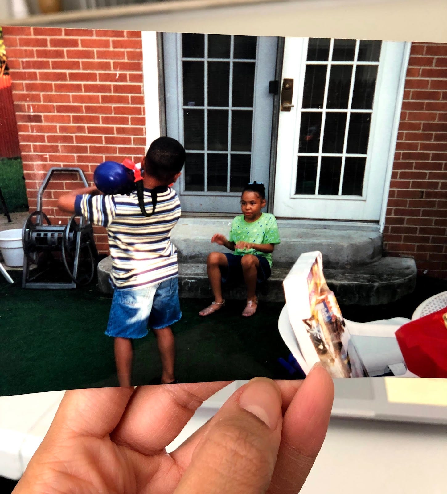An old picture being held with a little boy back to the camera, pointing a super soaker water gun at a little girl sitting outside on a porch step of a brick house.