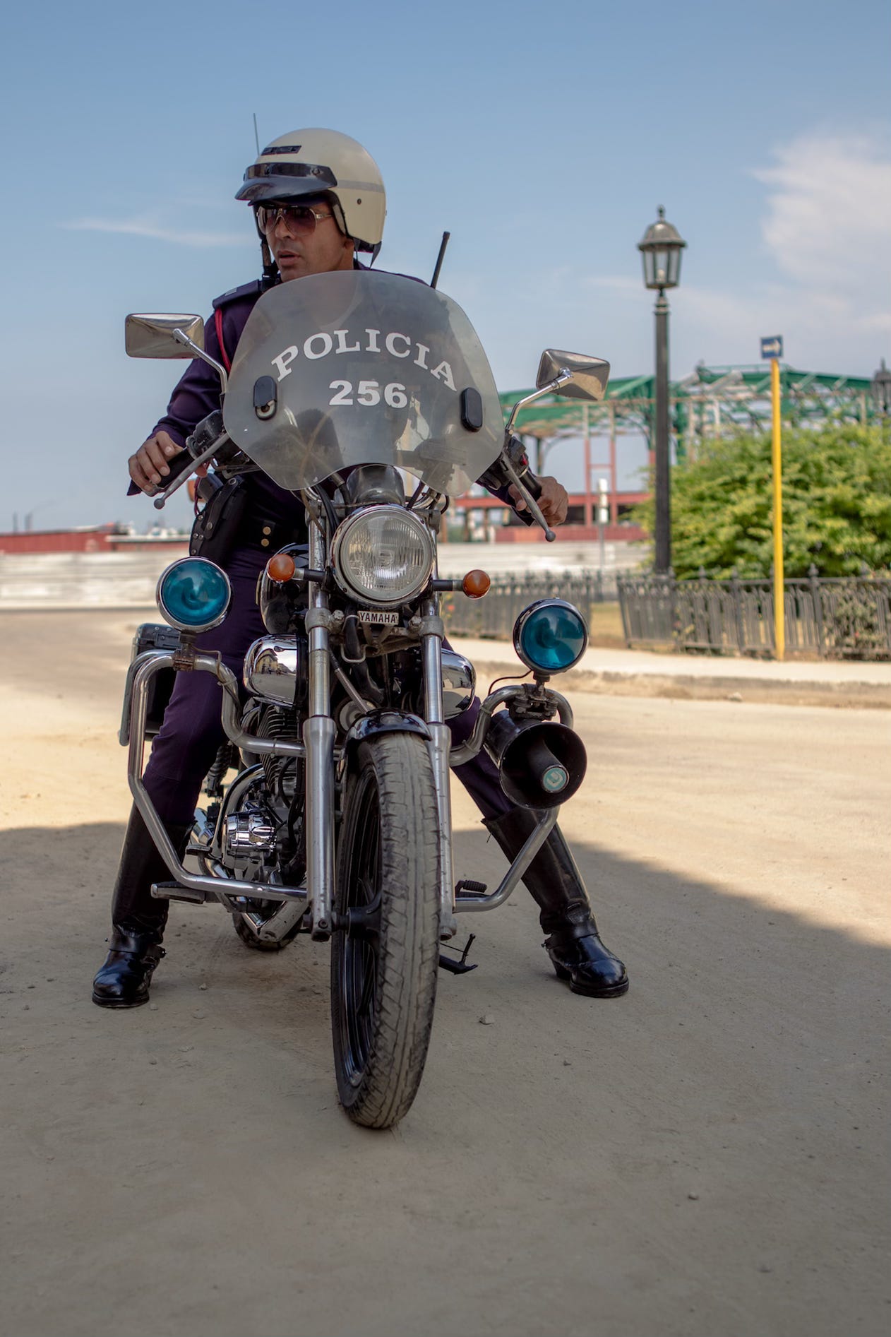 A Cuban police officer on a Yamaha motorcycle faces the camera wearing a helmet and mirrored sunglasses. The bike's windscreen reads “Policia 256.” Shot from a low angle on a dusty road, the image was taken candidly, capturing the quiet intensity of the moment.