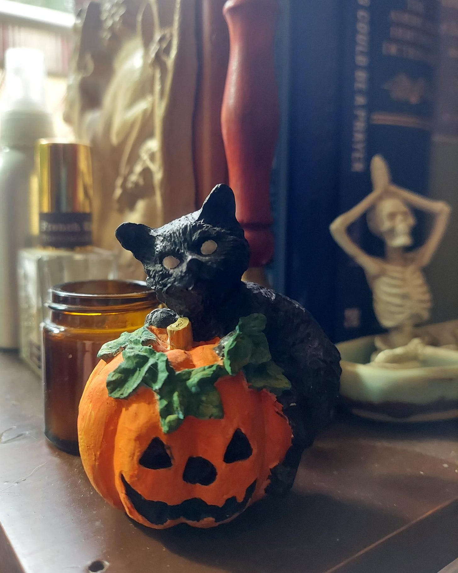 A ceramic black cat on a pumpkin. The ornaments are on a bookshelf. A skeleton sits nearby in the lotus position serving as a bookend A ceramic black cat on a pumpkin. The ornaments are on a bookshelf. A skeleton sits nearby in the lotus position serving as a bookend