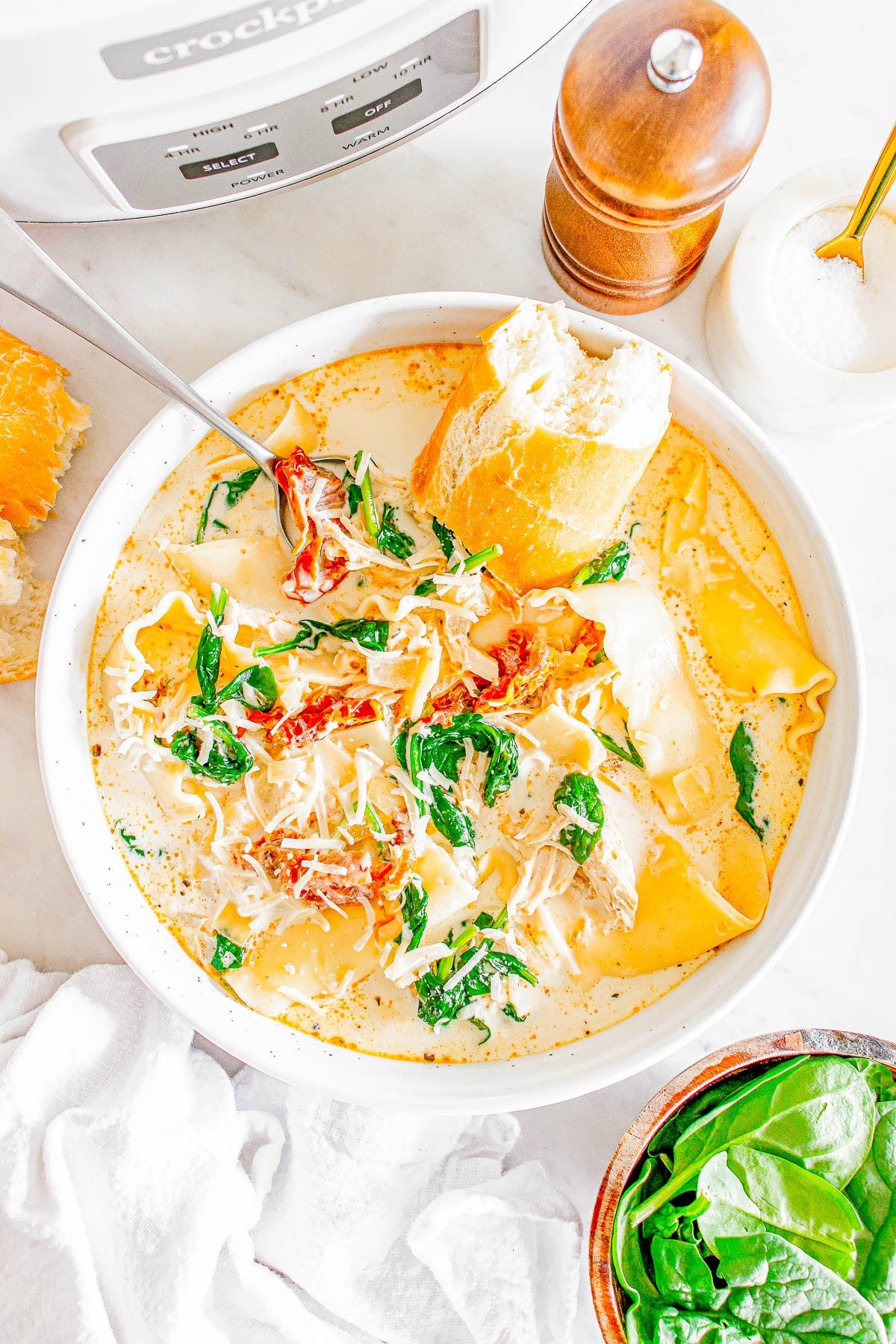 Bowl of creamy soup with spinach, shredded cheese, lasagna noodles, and chicken, garnished with sun-dried tomatoes and served with a piece of bread. Crockpot and pepper mill in the background. Bowl of creamy soup with spinach, shredded cheese, lasagna noodles, and chicken, garnished with sun-dried tomatoes and served with a piece of bread. Crockpot and pepper mill in the background.