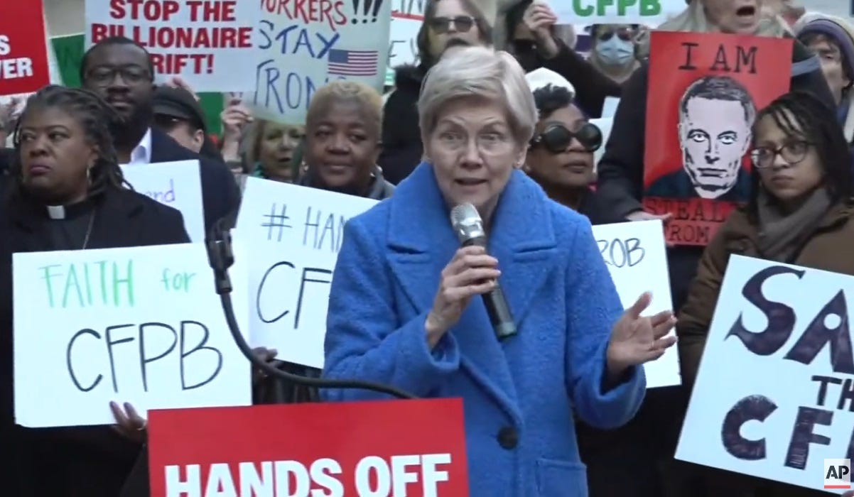 Video screenshot of Elizabeth Warren, in a blue cloth coat and holding a microphone, speaking to demonstrators who carry signs like 'Stop the billionaire grift,' 'Hands off CFPB,' and a big-brother-esque picture of Elon Musk with the words 'I Am Stealing From You'  
