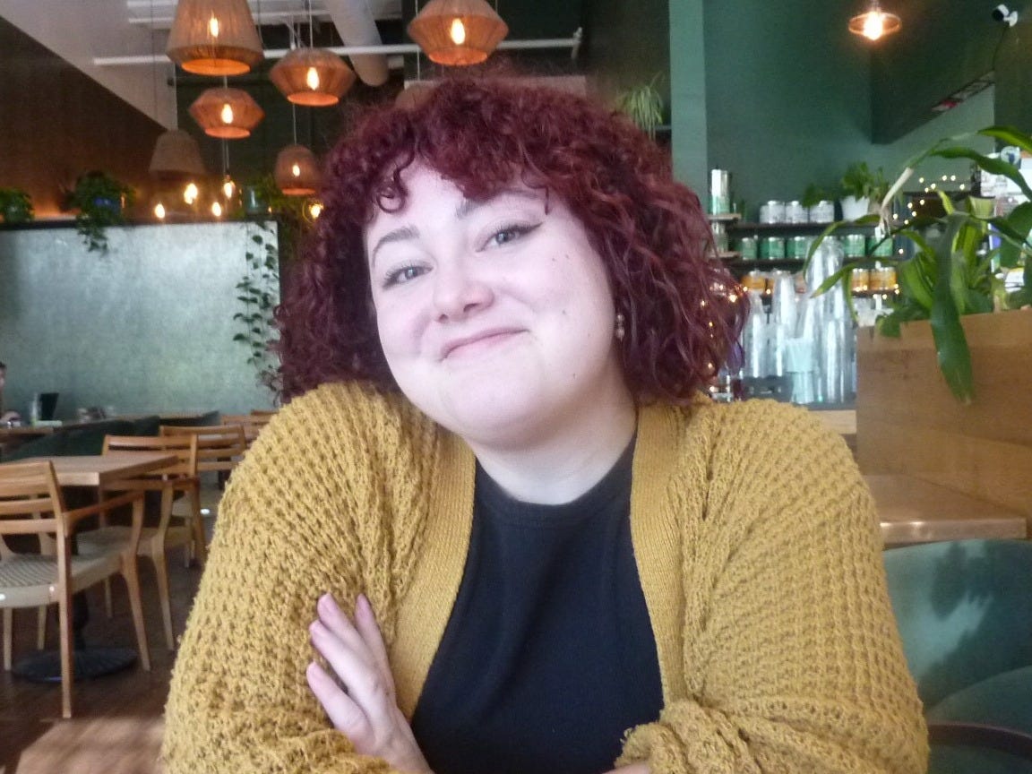 A young white person with curly, shoulder-length red hair smiles at the camera. They are wearing a mustard-yellow cardigan and a black shirt, and they appear to be seated at a table in a bright coffee shop, which can be seen in the background.