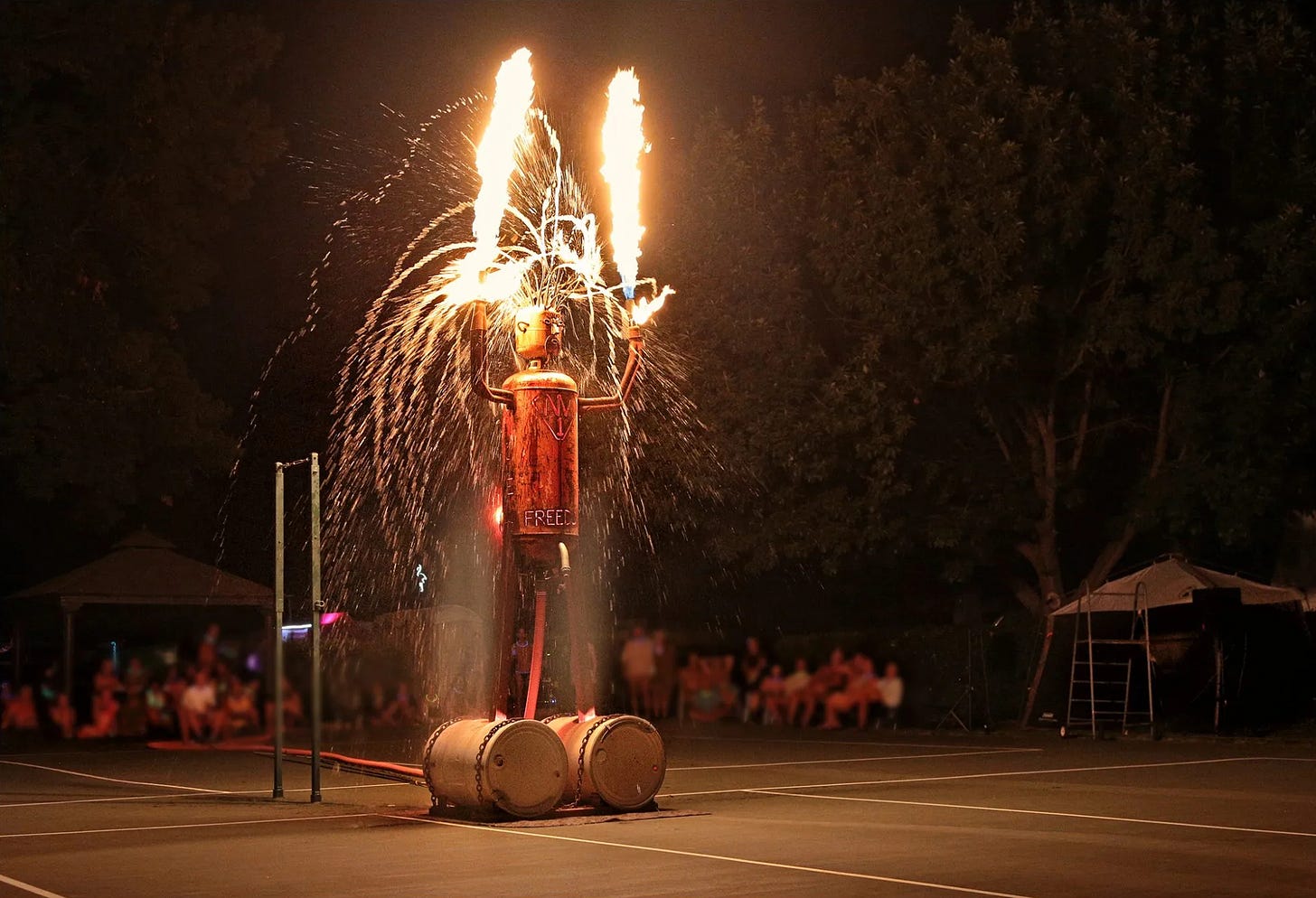 A large humanoid metal sculpture with arms raised shoots flames and sparks into the night sky during the Naked Man event at Laguna del Sol, as a crowd watches in the background. A large humanoid metal sculpture with arms raised shoots flames and sparks into the night sky during the Naked Man event at Laguna del Sol, as a crowd watches in the background.