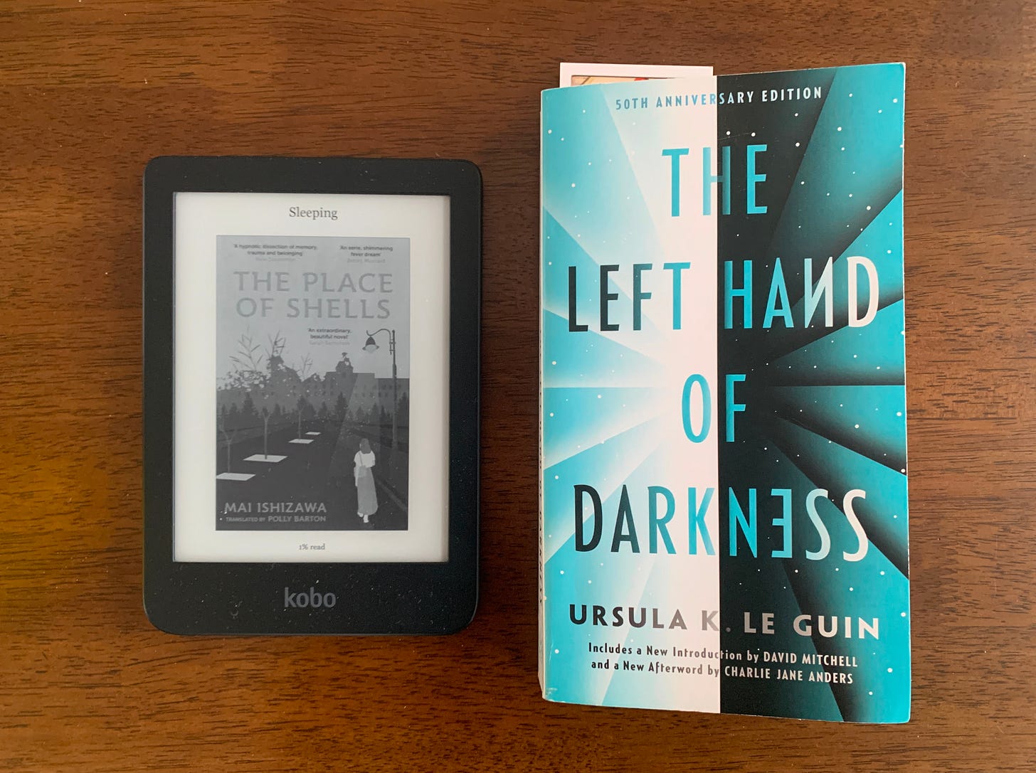 an ereader with the place of shells on it and a paperback copy of the left hand of darkness sit on a wooden table