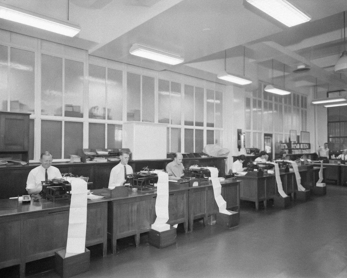 Vintage black and white photo of men in shirts and ties sitting at a row of office desks working on office machines. Reams of paper are flowing out of their machines, over the desks, and onto the floor. 