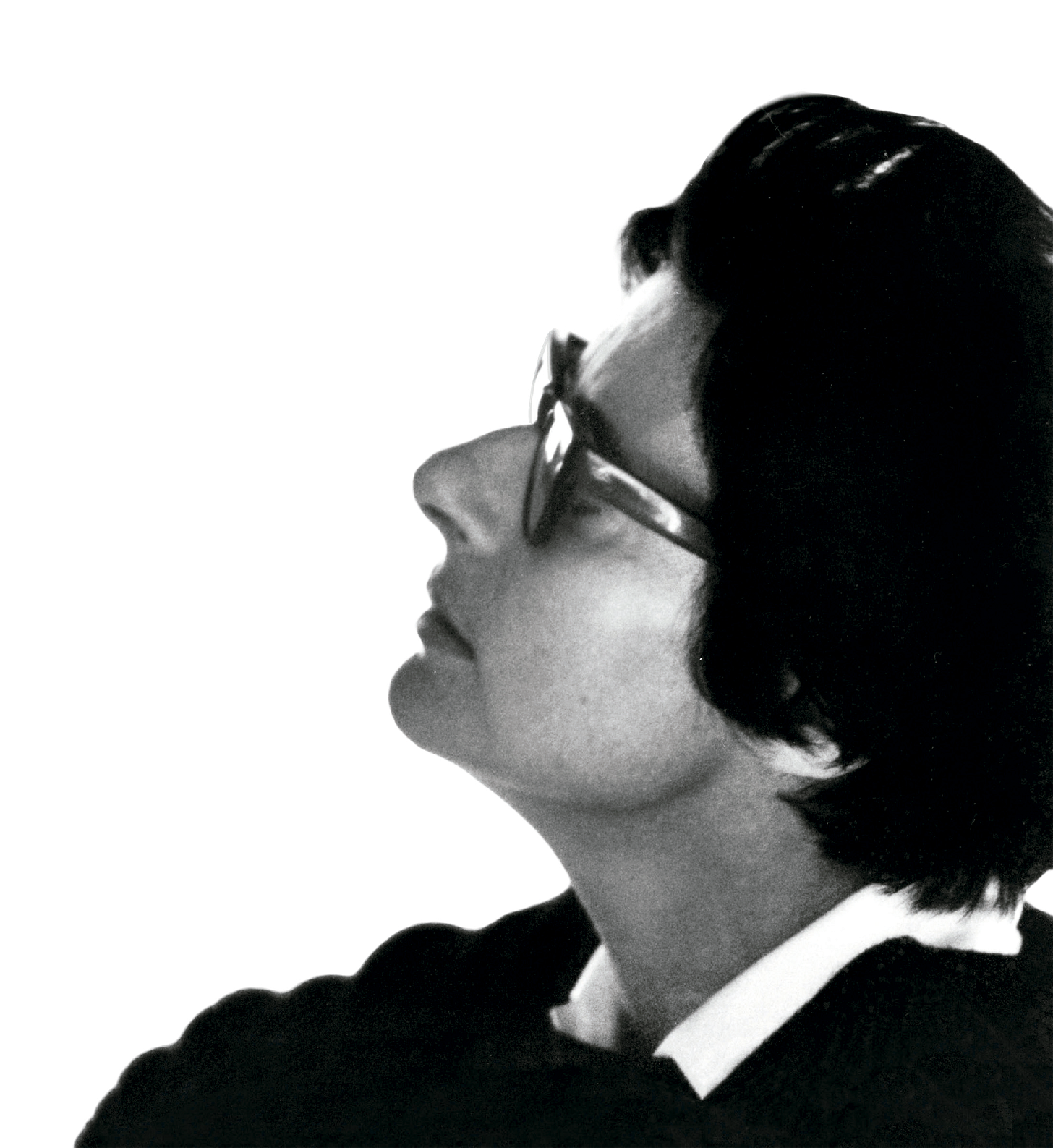 Fotografia em preto e branco de perfil de uma pessoa com cabelo curto escuro, usando óculos de armação clara e vestindo uma peça de roupa escura com gola clara, com o rosto voltado para cima em direção à luz.