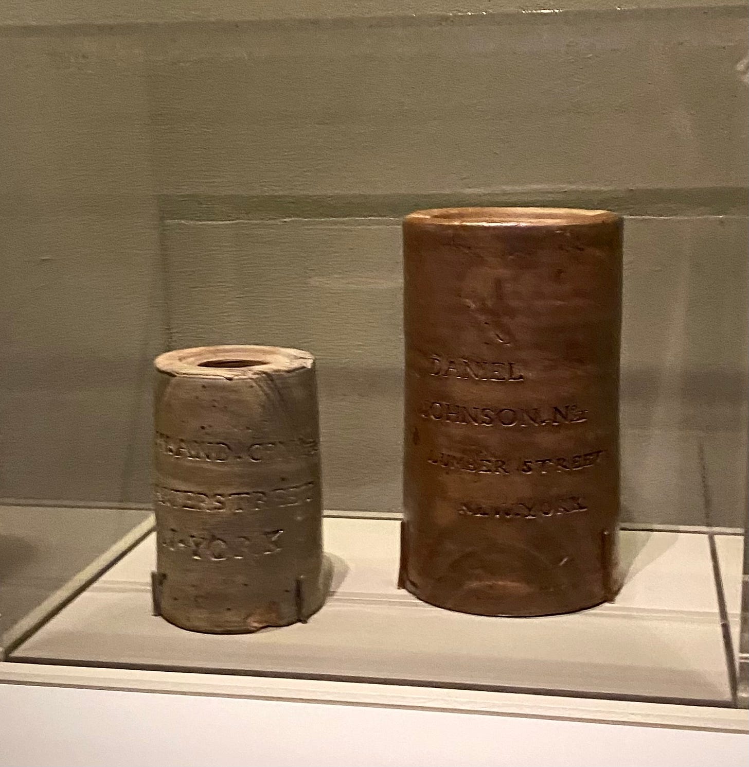 Two ceramic, cylindrical jars in a museum case. One is inscribed DANIEL JOHNSON LUMBER STREET.
