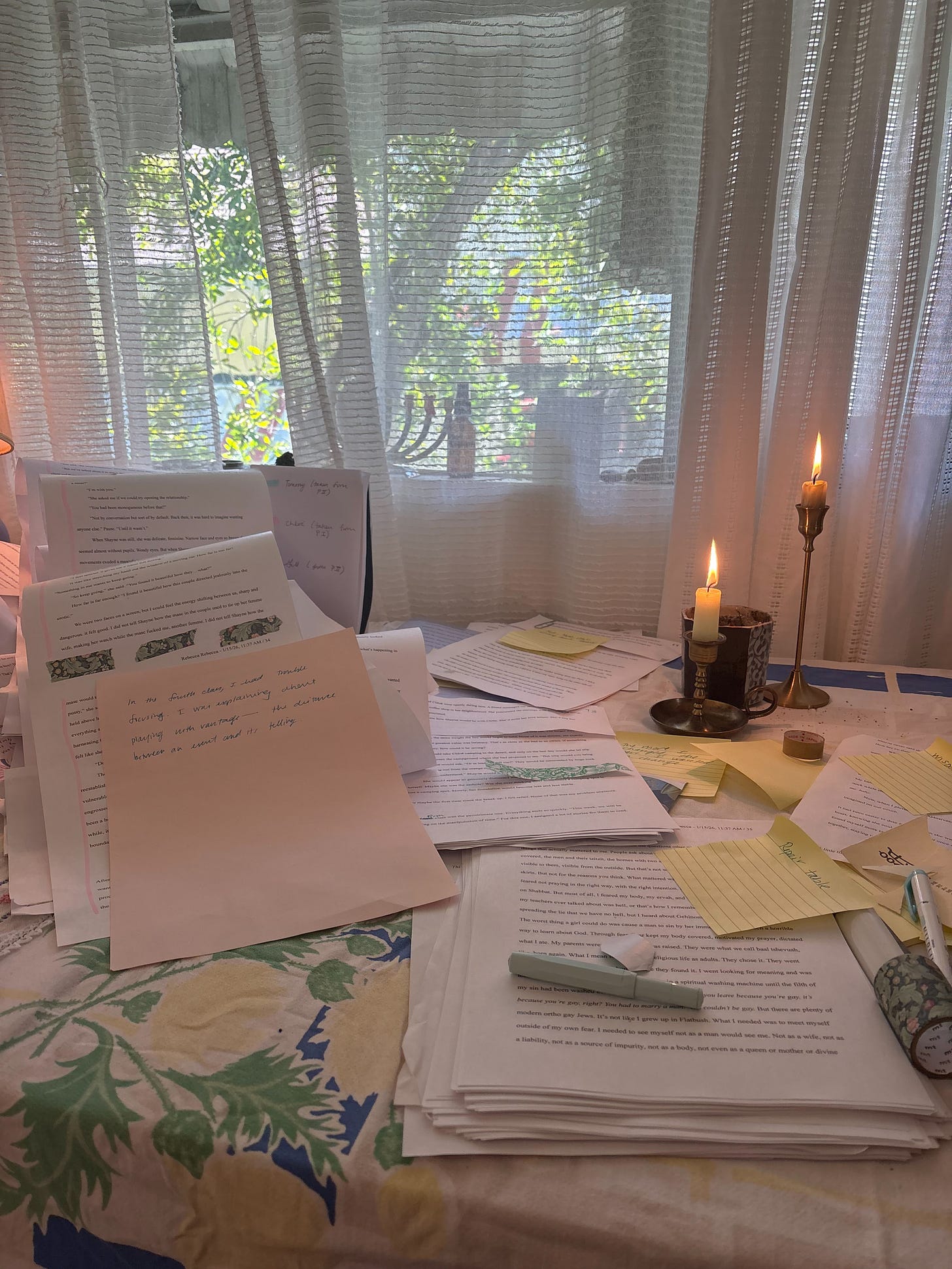 A table with a manuscript, lit candles, and various pens. Behind the table, a window with sheer curtains, daylight.
