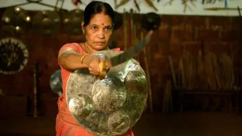 Meenakshi Raghavan A woman wearing an orange saree stands with a brass shield and a sword 